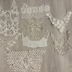May include: An assortment of off-white lace collars and embellishments, featuring various floral and geometric patterns. The collection includes several different styles, from delicate necklines to broader, decorative pieces, all with intricate openwork designs.