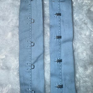 May include: Two light blue fabric strips with metal hook and eye closures. One strip has five D-shaped hooks, and the other has five eye closures. The strips are sewn with white thread and are likely for garment construction.