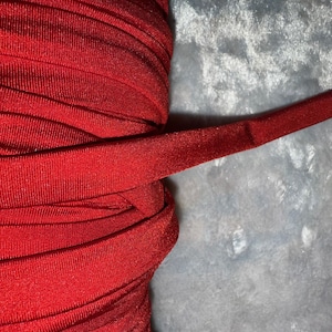 May include: Close-up of a roll of vibrant red fabric, possibly bias tape or binding. The material is tightly wound, with a smooth, slightly shiny texture. A length of the fabric extends outward, showing its finished edge.