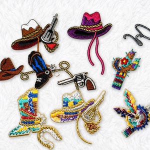 May include: A collection of embroidered patches featuring Western-themed designs including cowboy hats, boots, revolvers, a cactus, a lasso, and a colorful star-shaped badge. The patches display various colors such as brown, pink, blue, and yellow.