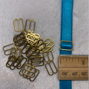 May include: A pile of gold-colored metal bra strap sliders, with a turquoise blue elastic bra strap and a wooden ruler marked with inches. The ruler shows measurements from 1/8 to 3/10 of an inch.