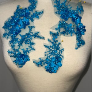 May include: Two turquoise blue floral appliques with sequins and beads on a white fabric background.