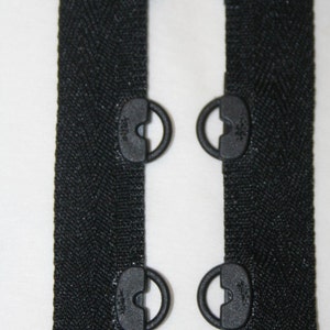 May include: Close-up of black zipper tape with attached black metal eyelets. The zipper tape has a herringbone weave pattern. The eyelets are evenly spaced and appear to be for attaching the zipper to fabric.