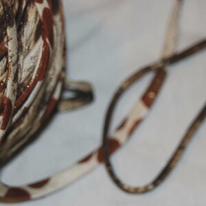 May include: A spool of decorative ribbon with a mix of brown, cream, and gold colors. The ribbon has a patterned design with a contrasting cream and brown spotted cord extending from the spool.
