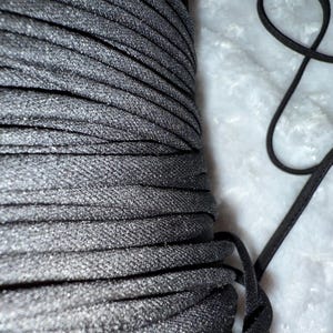 May include: A spool of black elastic cord, tightly wound, with a textured surface. A length of the same cord lies loose, showing its flexibility. The cord is suitable for various crafting and sewing projects.