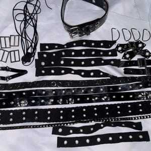 May include: A collection of black leather and metal hardware components, including belts, straps, buckles, and grommets, arranged on a white surface. The items appear to be for crafting or fashion accessories.