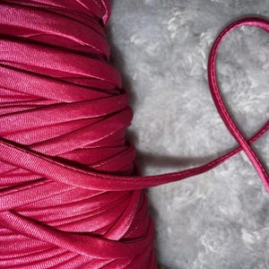 May include: Close-up of a spool of bright pink satin ribbon. The ribbon is tightly wound, with a single loop of ribbon extending out. The background is a soft, textured, light gray surface. The ribbon appears smooth and shiny.