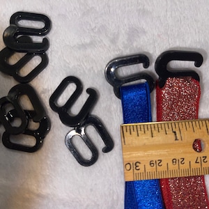 May include: A close-up of black plastic bra strap adjusters and two elastic straps, one blue and one red with glitter. A ruler is visible in the background with markings from 1/2 inch to 2 inches.