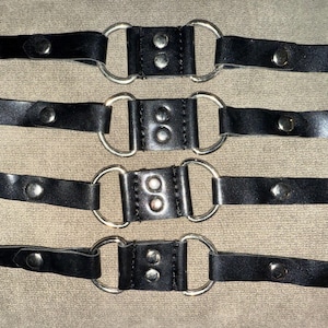 May include: Four black leather straps with silver metal rings and snap closures. Each strap has a pointed end and is secured with silver rivets. The straps are arranged horizontally against a neutral background.
