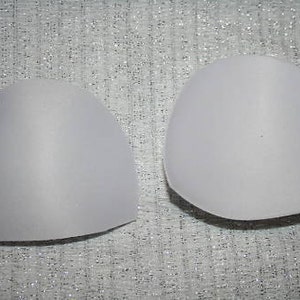 May include: Two white, semi-circular bra cups made of foam padding. The cups are designed to add volume and shape to the bust.