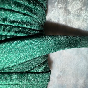 May include: Close-up of a roll of emerald green, sparkly fabric. The fabric is ribbed and has a metallic sheen, reflecting light. The material is tightly wound, with a section extending outward. The background is a light, textured surface.