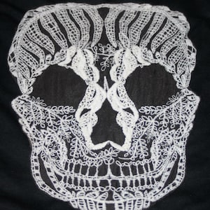 May include: A white lace skull design on a black background. The skull is intricately detailed with floral and geometric patterns, creating a striking contrast. The design is centered and fills the frame, showcasing the delicate lacework.