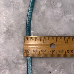 May include: A close-up of a light blue elastic cord measured against a wooden ruler. The ruler shows measurements in inches, with markings for 1/2, 1/4, 1/8, and 1/16 inches.