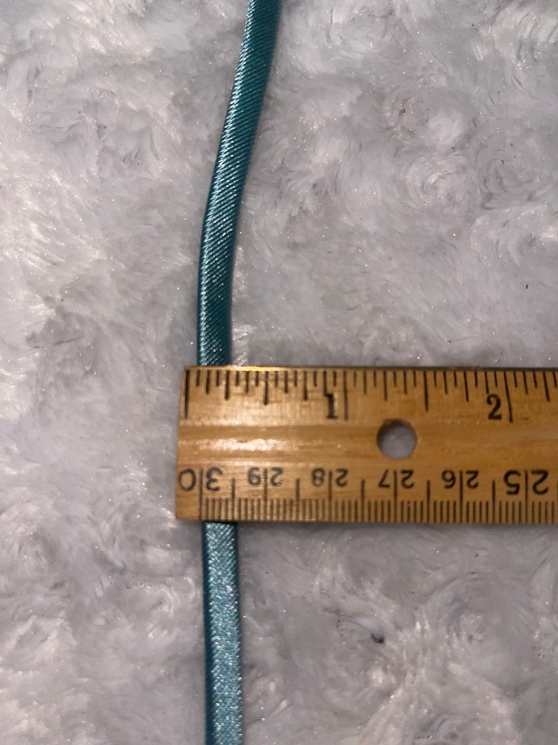 May include: A close-up of a light blue elastic cord measured against a wooden ruler. The ruler shows measurements in inches, with markings for 1/2, 1/4, 1/8, and 1/16 inches.