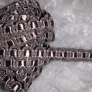 May include: A roll of decorative trim featuring a repeating pattern of black and silver loops. The trim has a scalloped edge and a shiny finish, perfect for adding a touch of elegance to various craft projects. The trim is shown against a soft, light blue background.