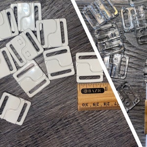 May include: Two sets of white plastic bra clasps, one set of clear plastic bra clasps, and a white ribbon. The white clasps are arranged in a pile on a wooden surface. The clear clasps are scattered on a wooden surface. The white ribbon is lying on a wooden ruler with inch markings.