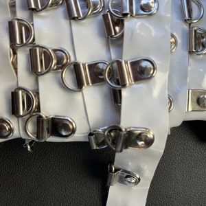 May include: White plastic webbing with silver metal rings and tabs. The webbing is folded and the rings are visible on both sides.