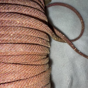 May include: Close-up of a spool of shimmering, rose gold-colored trim. The trim is tightly wound, revealing a textured surface with subtle glitter accents. A length of the trim extends from the spool, showcasing its flexible, cord-like structure.