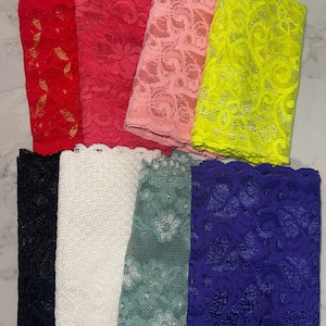 May include: A collection of folded lace fabric in various colors. The colors include red, pink, yellow, black, white, teal, and blue. The lace has a floral pattern and a scalloped edge.