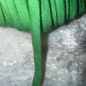 May include: Close-up of a roll of bright green bias tape. The fabric has a diagonal weave pattern and is unrolled slightly, revealing its texture. The tape is set against a soft, white, fuzzy background.