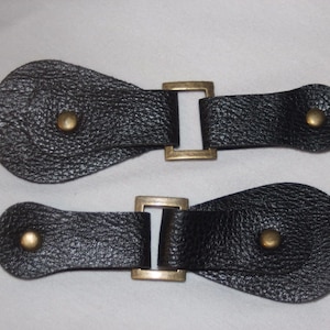 May include: Two black leather toggles with brass buckles. Each toggle has a round leather loop with a brass button.