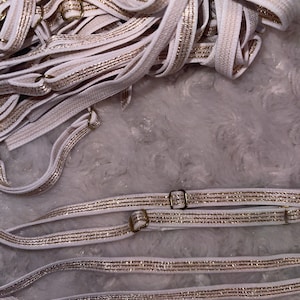 May include: A pile of white elastic straps with gold glitter trim. The straps have metal clasps.