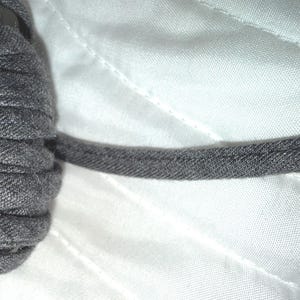 May include: Close-up of a roll of gray fabric trim, tightly wound. The trim is a solid, medium gray color with a textured surface. A length of the trim extends from the roll, showing the edge stitching. The background is a white, quilted fabric.