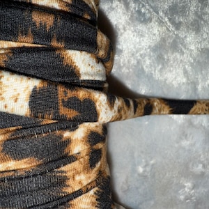 May include: Close-up of a fabric roll with a leopard print pattern. The fabric features a repeating design of black spots and brown patches on a beige background. The fabric is tightly rolled, with a thin, extended piece visible.