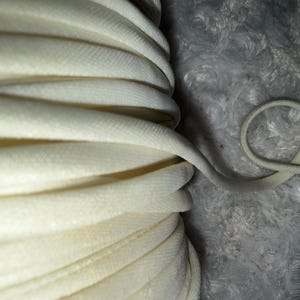 May include: Close-up of a spool of off-white, woven cord. The cord is tightly wound, with a single strand extending and looped on a textured, gray surface. The cord appears to be made of a soft, flexible material.