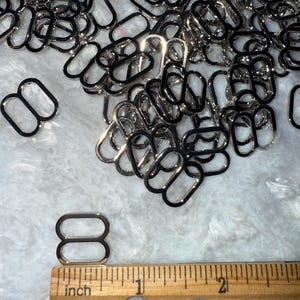 May include: A collection of shiny, silver-colored metal rectangle rings, possibly for use in crafting or jewelry making. A wooden ruler with inch measurements is in the foreground, showing the size of the rings. The rings are approximately 1 inch in length.
