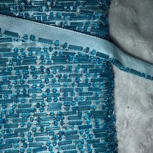 May include: Close-up of a roll of turquoise beaded trim with a matching ribbon. The trim features a dense pattern of small, rectangular beads, creating a textured surface. The ribbon is a lighter shade of turquoise and is sewn along the edge of the beaded trim.