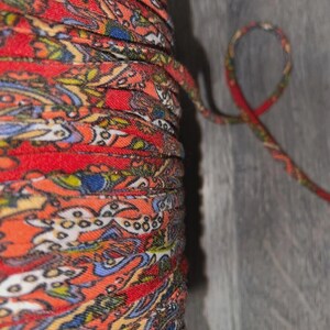 May include: Close-up of a spool of patterned fabric trim. The trim is a vibrant mix of red, orange, blue, and yellow with a paisley-like design. A length of the trim extends from the spool, forming a loop.