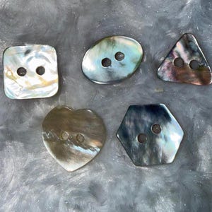 May include: Five iridescent buttons in various shapes: square, oval, triangle, heart, and hexagon. Each button has two holes for sewing. The buttons have a pearlescent sheen with colors ranging from white to gray and hints of blue and purple.