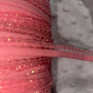 May include: A spool of pink elastic ribbon with gold glitter accents. The ribbon is tightly wound, with a thin, finished edge of the same pink color. The background is a soft, white, textured fabric.