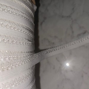 May include: Close-up of a roll of white elastic trim. The trim has a delicate, scalloped edge and is tightly wound. The material appears to be a soft, flexible fabric, suitable for various sewing and crafting projects.