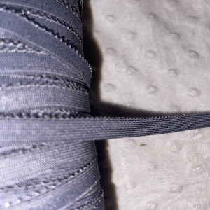 May include: A close-up of a spool of black elastic trim with a zig-zag stitch. The trim is rolled up and a piece is extended out.