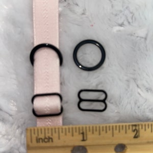 May include: A close-up of four black metal bra strap adjusters. The adjusters are on a white surface with a ruler marked in inches. The ruler shows measurements from 26 to 30 inches.