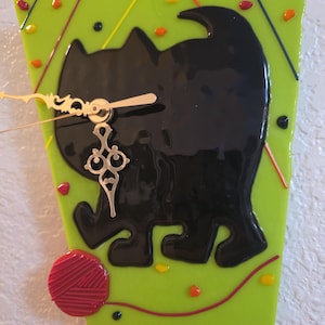 May include: A decorative wall clock featuring a black cat silhouette on a lime green background. The clock has gold-colored hands and a decorative pendulum. A red yarn ball and colorful accents complete the design. The clock is approximately 8 inches tall.