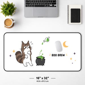 Puede incluir: Una alfombrilla de escritorio rectangular blanca de 40,6 cm por 81,3 cm, con un gato marrón y blanco mirando un caldero negro con líquido verde burbujeante. Una botella pequeña vierte líquido verde en la olla. El texto BOO BREW está impreso cerca.