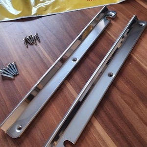 Könnte beinhalten: Zwei silberne Metallstreifen mit abgerundeten Kanten und Löchern für Schrauben. Die Streifen liegen auf einer Holzoberfläche mit einem Schraubenhaufen links.