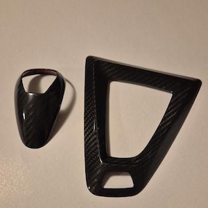 May include: Two black carbon fibre car interior trim pieces. One is a gear shift knob, and the other is a trim piece with a triangular shape. Both pieces have a glossy finish and a woven carbon fibre pattern.