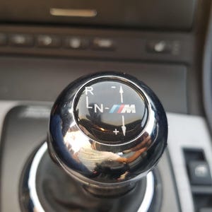 May include: Close-up of a silver gear shift knob with a black faceplate. The faceplate has a black and white "M" logo with blue and red accents. The gear shift knob has markings for "R", "L", "N", and "M".
