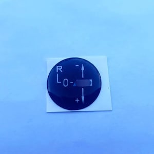Op de afbeelding: Een zwarte, cirkelvormige sticker voor het bedieningspaneel met witte tekst en symbolen. De sticker heeft de letters "R" en "L0-" samen met een plus- en minteken. De sticker zit op een witte, rechthoekige achterkant.