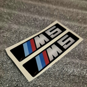 Może przedstawiać: Dwie czarne prostokątne naklejki z literami "M5" w kolorze srebrnym. Każda naklejka ma niebieski, czerwony i jasnoniebieski pasek. Naklejki są na białym podkładzie.