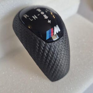 Op de afbeelding: Zwarte versnellingspookknop met een carbonfiberpatroon en een blauw-wit BMW-logo. De knop heeft een zwarte kunststof bovenkant met de letters "P, R, N, -1, 0, +" erop gedrukt.