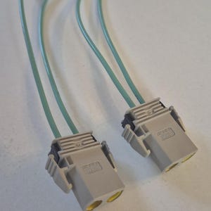 Może przedstawiać: Dwa szare złącza elektryczne z jasnozielonymi przewodami. Każde złącze ma dwa żółte otwory i tekst "HELLA" wydrukowany z boku. Przewody są elastyczne i wydają się być wykonane z tworzywa sztucznego.