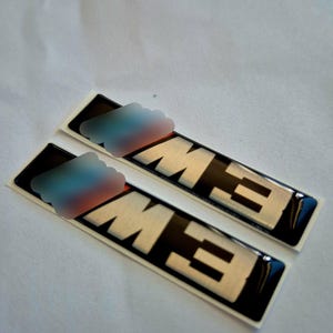 Puede incluir: Dos pegatinas con el emblema M3 en negro y plata. Las pegatinas tienen forma rectangular con las letras "M3" en plata sobre un fondo negro. Las pegatinas están sobre una superficie blanca.