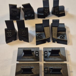 Peut inclure: Ensemble de pièces en plastique noir imprimées en 3D, chacune avec des formes et des textes uniques. Les pièces comprennent des éléments angulaires avec des inscriptions telles que "RB" et "BC", et des pièces rectangulaires avec "BD". Les objets sont disposés sur une surface claire.