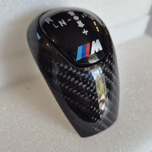 Könnte beinhalten: Ein schwarzer Carbonfaser-Schalthebelknauf mit einem schwarz-weißen Schaltmuster und einem blau-rot-weißen BMW-Logo. Der Knauf hat ein schwarz-weißes Carbonfaser-Muster.