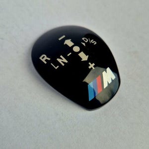 Puede incluir: Pomo de palanca de cambios de plástico negro con letras blancas para los símbolos 'R', 'L', 'N', 'D', 'P', 'S' y '+'. El pomo tiene un logotipo BMW azul, rojo y blanco.